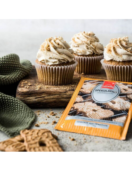Drei Muffins mit Cremehaube und Streuseln neben gelbem Flachbeutel auf Holzplatte.