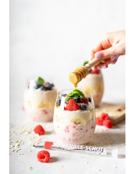 Dessert im Glas mit Beeren, Minze und Honig, im Vordergrund Vanilleschote in transparenter Hartkorn-Röhre mit rotem Etikett Dessert im Glas mit Beeren, Minze und Honig, im Vordergrund Vanilleschote in transparenter Hartkorn-Röhre mit rotem Etikett