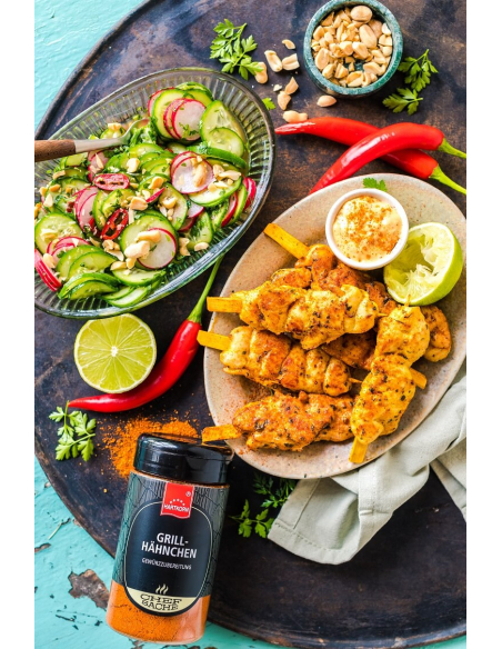 Gurkensalat mit Radieschen und Erdnüssen in Glasschale, daneben Chilischoten und Kräuter auf dunklem Untergrund.