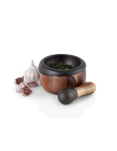 Ein Mörser mit Stößel aus dunklem Stein und Holz liegt neben Knoblauch und Kräutern auf weißem Untergrund.