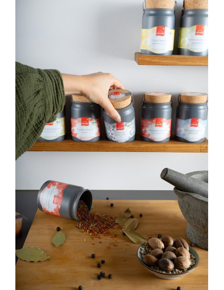 Hand greift ein Keramiktöpfchen aus dem Regal, vorne liegt ein geöffnetes Töpfchen mit verstreutem Gewürzinhalt.