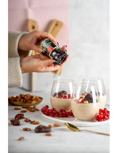 Person würzt Dessert mit einer Gewürzmühle; auf dem Tisch Gläser mit Creme, Johannisbeeren und Nüssen.