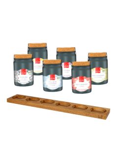 Sechs schwarze Keramiktöpfchen mit Korkdeckel hinter einem langen Holzbrett mit sechs runden Aussparungen.