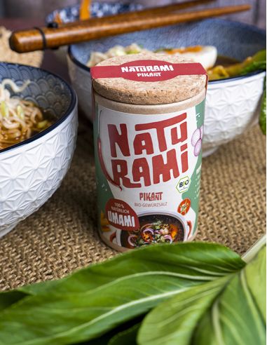 Schwarze Pappdose mit rotem Etikett mit der Aufschrift NATURAMI, daneben Schüsseln mit Ramen und Essstäbchen.