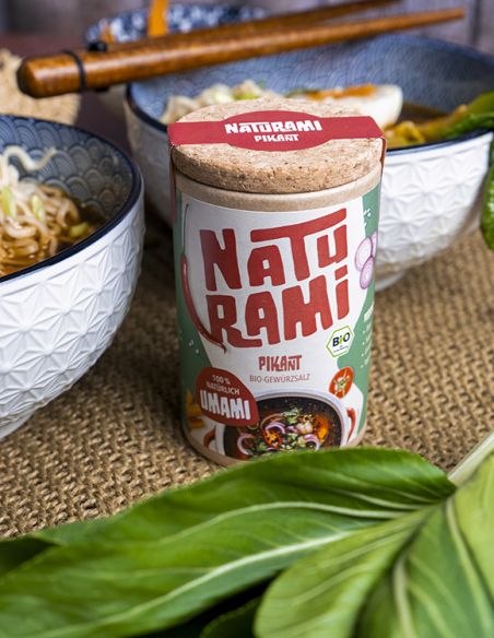 Schwarze Pappdose mit rotem Etikett mit der Aufschrift NATURAMI, daneben Schüsseln mit Ramen und Essstäbchen.