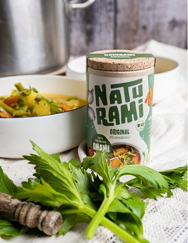 Schwarze Pappdose mit grünem Etikett mit der Aufschrift NATURAMI vor einer weißen Schale mit Gemüsesuppe und frischem Sellerie.