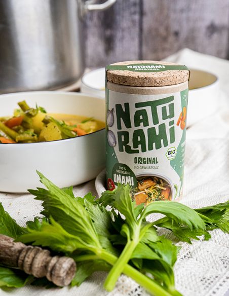 Schwarze Pappdose mit grünem Etikett mit der Aufschrift NATURAMI vor einer weißen Schale mit Gemüsesuppe und frischem Sellerie.