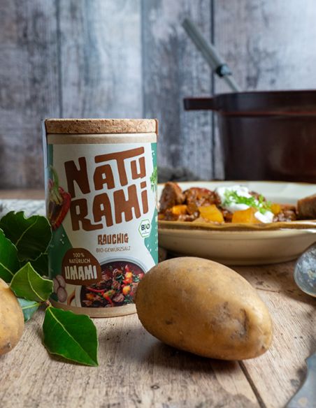Schwarze Pappdose mit hellblauem Etikett mit der Aufschrift NATURAMI, daneben Kartoffeln und Lorbeerblätter auf einem Holztisch