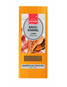 Flachbeutel mit der Aufschrift „KREUZKÜMMEL 'Cumin' Gemahlen“, dahinter Holzlöffel und kariertes Tuch.