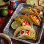 Vier gefüllte Tacos mit Bohnencreme, Avocado, Cherrytomaten, Joghurt und Koriander auf einem Tablett – garniert mit Avocado-Topping und Limette