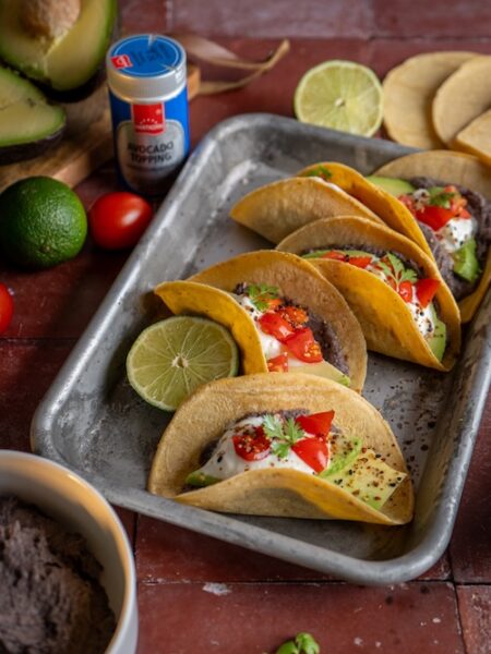 Vier gefüllte Tacos mit Bohnencreme, Avocado, Cherrytomaten, Joghurt und Koriander auf einem Tablett – garniert mit Avocado-Topping und Limette