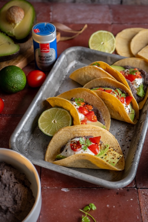 Vier gefüllte Tacos mit Bohnencreme, Avocado, Cherrytomaten, Joghurt und Koriander auf einem Tablett – garniert mit Avocado-Topping und Limette