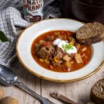 Eine rötliche Gulaschsuppe, getroppt mit Crème fraîche und einem Stück Brot. Im Hintergrund steht eine Naturami Rauchig Dose.