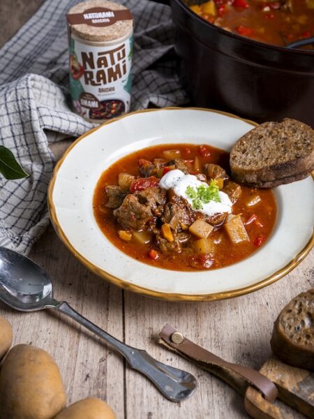 Eine rötliche Gulaschsuppe, getroppt mit Crème fraîche und einem Stück Brot. Im Hintergrund steht eine Naturami Rauchig Dose.