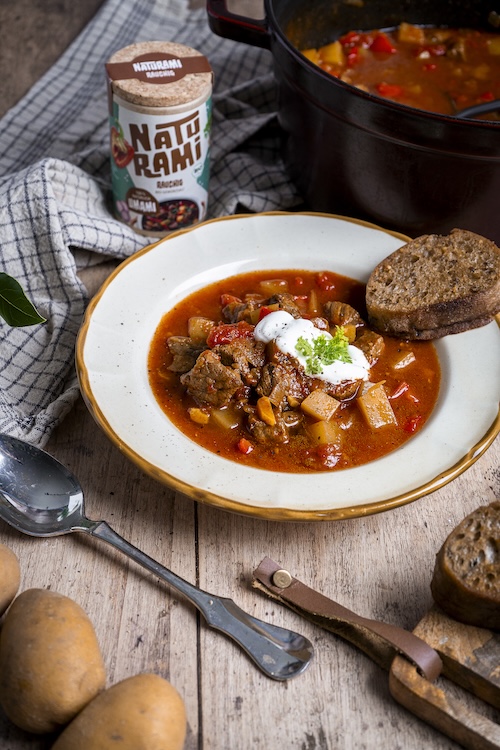 Eine rötliche Gulaschsuppe, getroppt mit Crème fraîche und einem Stück Brot. Im Hintergrund steht eine Naturami Rauchig Dose.