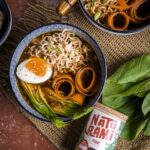 Eine dunkle Schüssel mit Ramen, einem Ei und verschiedenen Einlagen. Daneben liegt eine Naturamidose in der Sorte pikant.