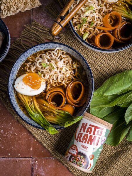 Eine dunkle Schüssel mit Ramen, einem Ei und verschiedenen Einlagen. Daneben liegt eine Naturamidose in der Sorte pikant.