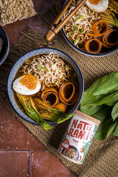 Eine dunkle Schüssel mit Ramen, einem Ei und verschiedenen Einlagen. Daneben liegt eine Naturamidose in der Sorte pikant.