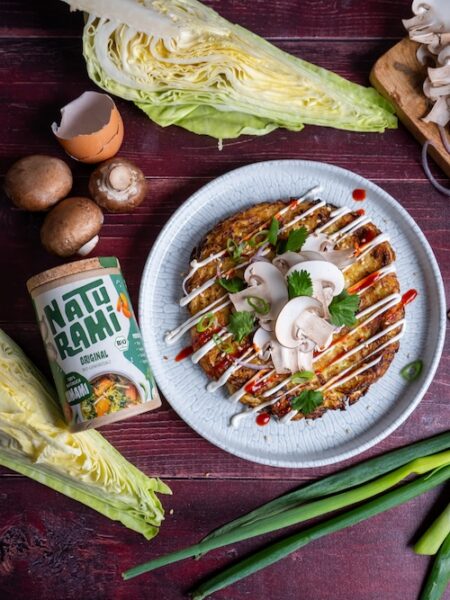 Ein vegetarisches Okonomiyaki auf einem Teller, getoppt mit Sesammayonnaise, Srirachasoße, Frühlingszwiebeln und frischem Koriander