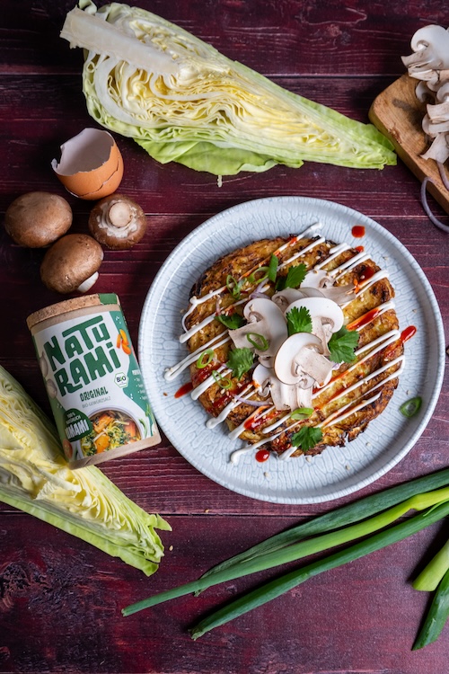 Ein vegetarisches Okonomiyaki auf einem Teller, getoppt mit Sesammayonnaise, Srirachasoße, Frühlingszwiebeln und frischem Koriander