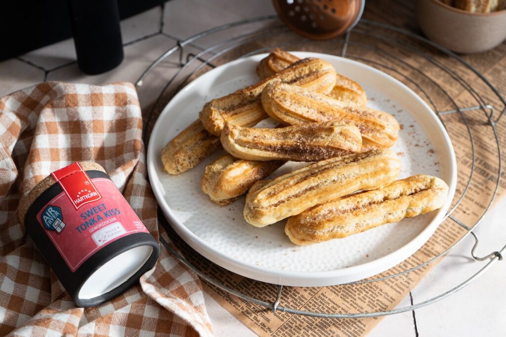 Goldbraune Churros aus der Heißluftfritteuse auf einem weißen Teller, daneben Sweet Tonka Kiss Gewürzzucker von Hartkorn.
