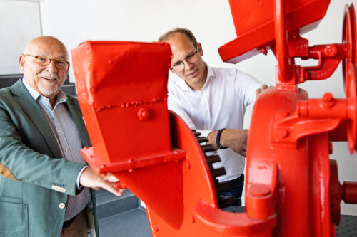 Zwei Männer begutachten eine große rote Industriemaschine und lächeln dabei. Zwei Männer begutachten eine große rote Industriemaschine und lächeln dabei.