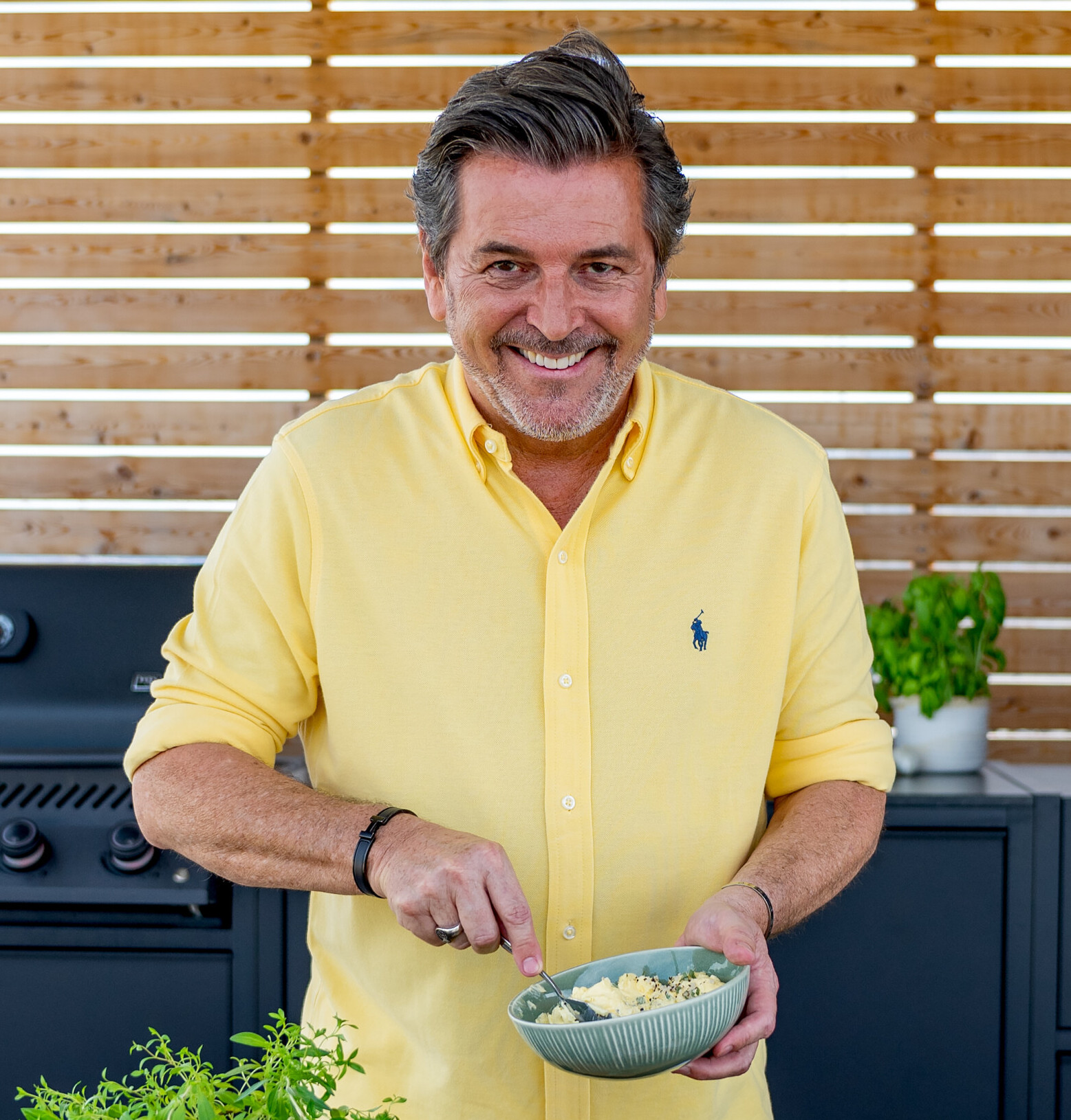 Thomas Anders Thomas Anders lächelt in gelbem Hemd und rührt in einer Schüssel, im Hintergrund Grill und Kräuter.